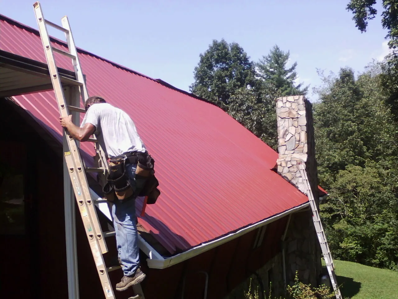 Roofer on a ladder beside a red metal roof and stone chimney — Soffit & Fascia Repair by Roofing Brothers in Idledale